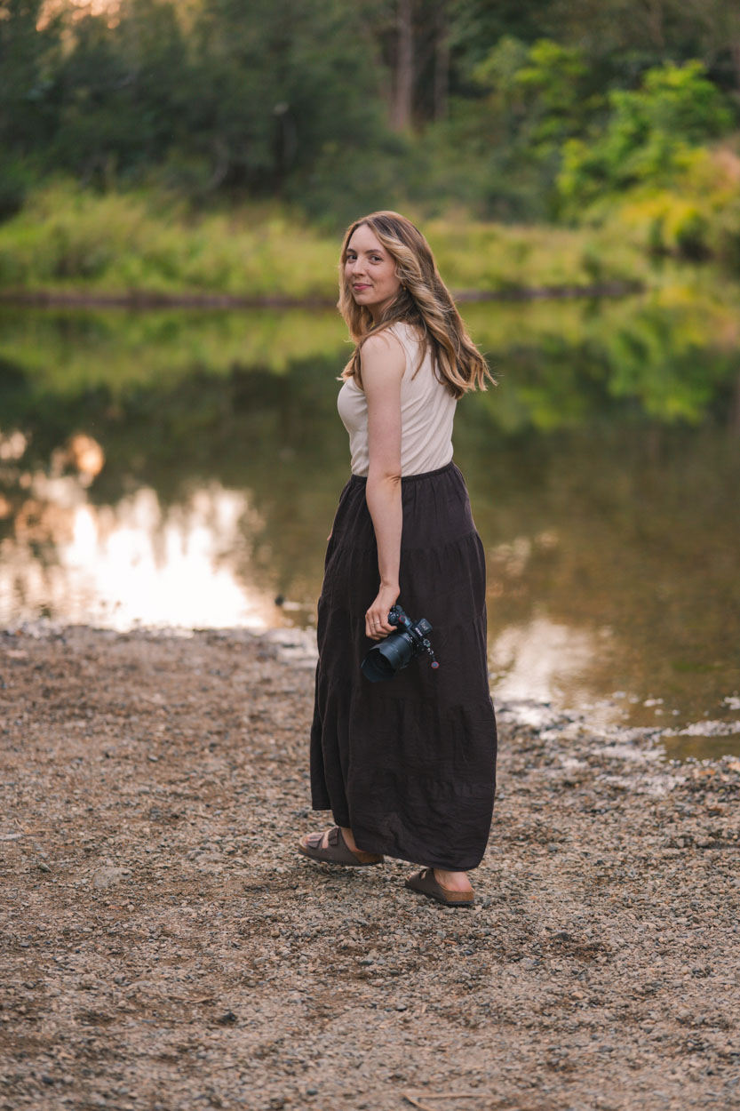 Katie standing by a still pond at golden hour with her camera
