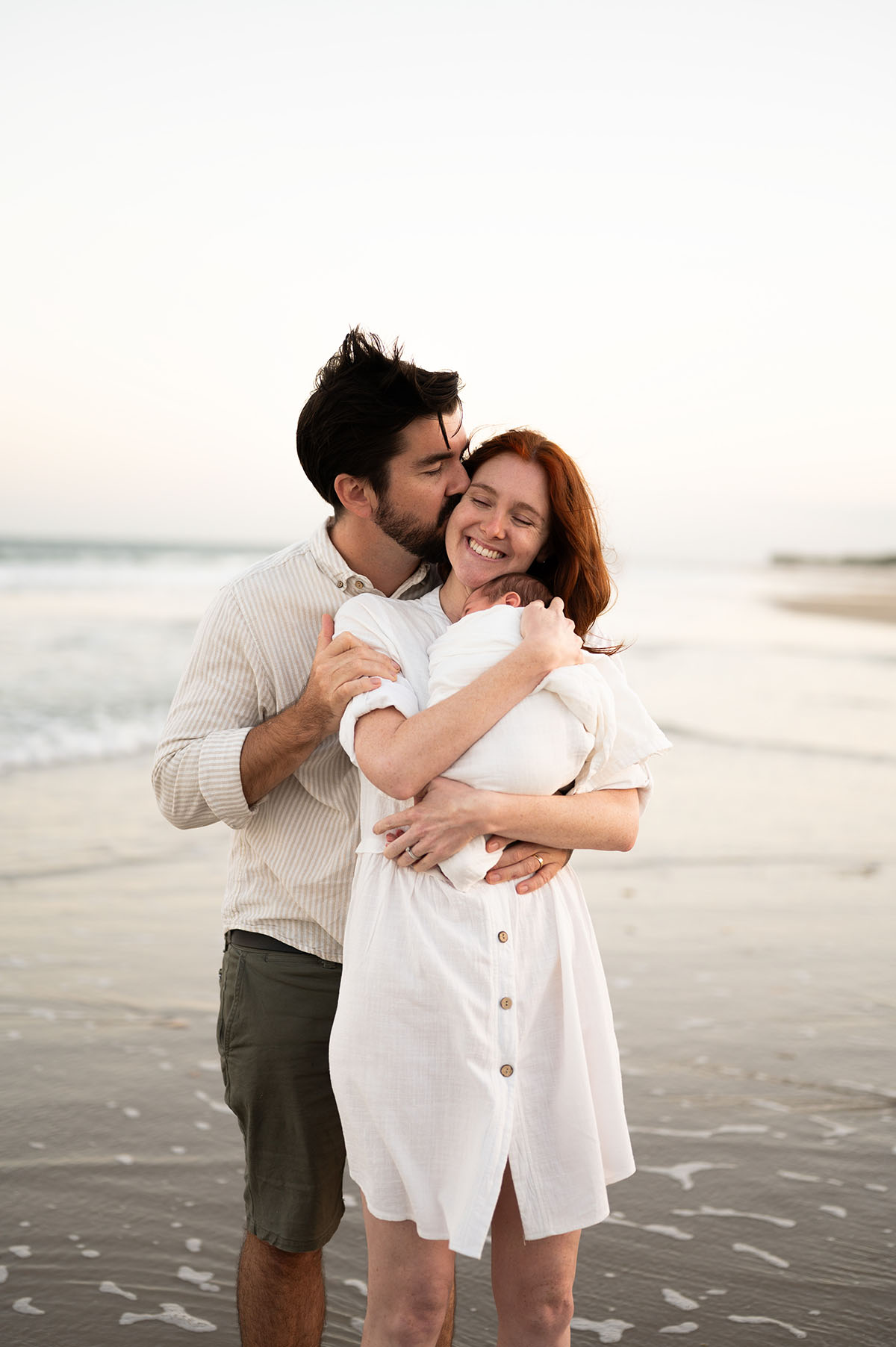A father kissing the mother's head as she smiles holding their newborn baby on the beach