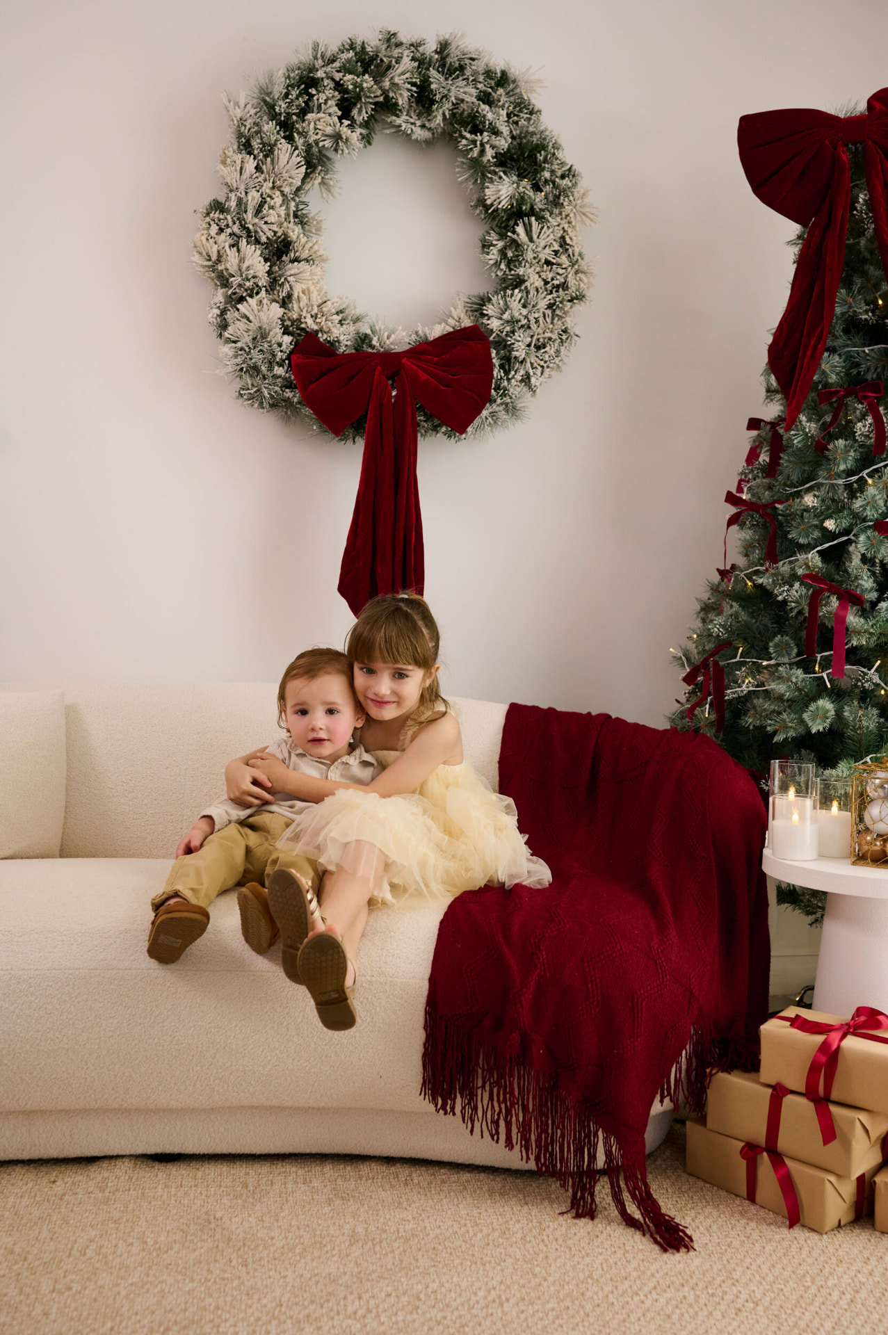 Two children sitting together on a cream couch beneath a frosted wreath with a deep red velvet bow