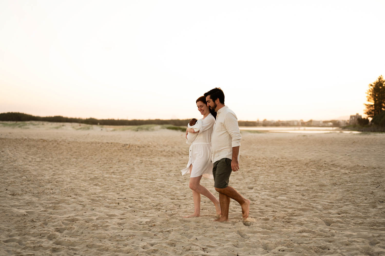 A couple walking barefoot along the beach at sunset as the mother holds their newborn baby