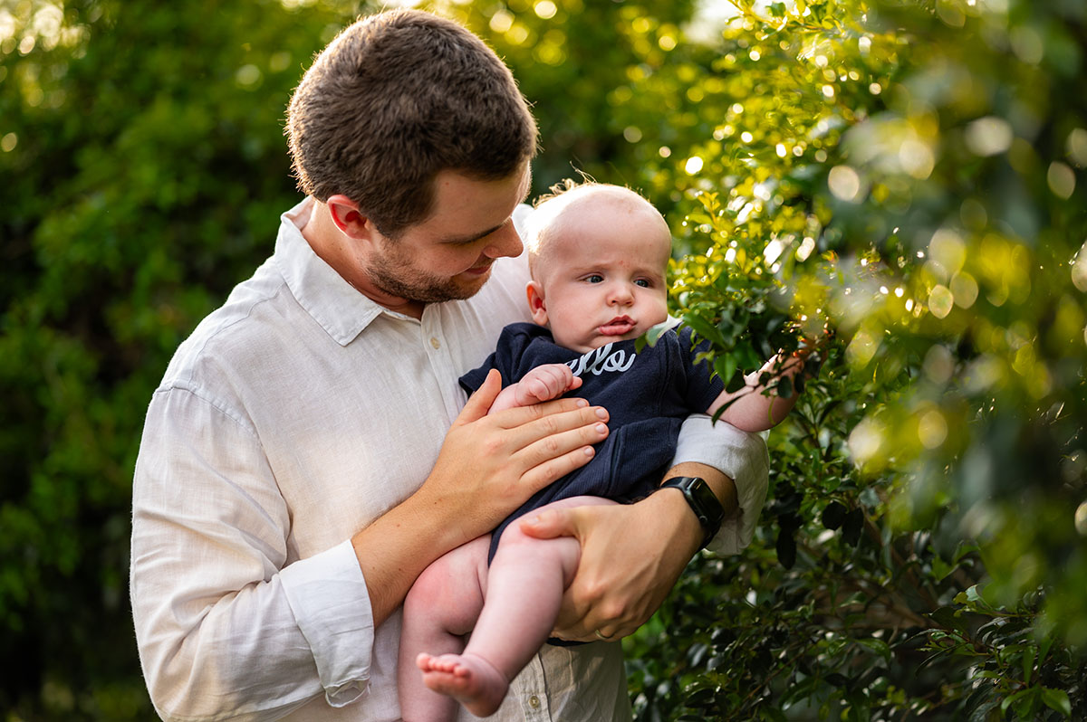 A father smiling down at his baby daughter as he holds her among sunlit greenery